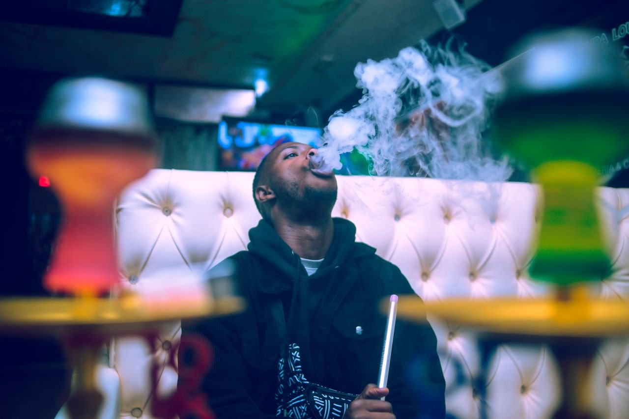 About A young man relaxing and smoking shisha at an indoor lounge, surrounded by colorful hookahs.
